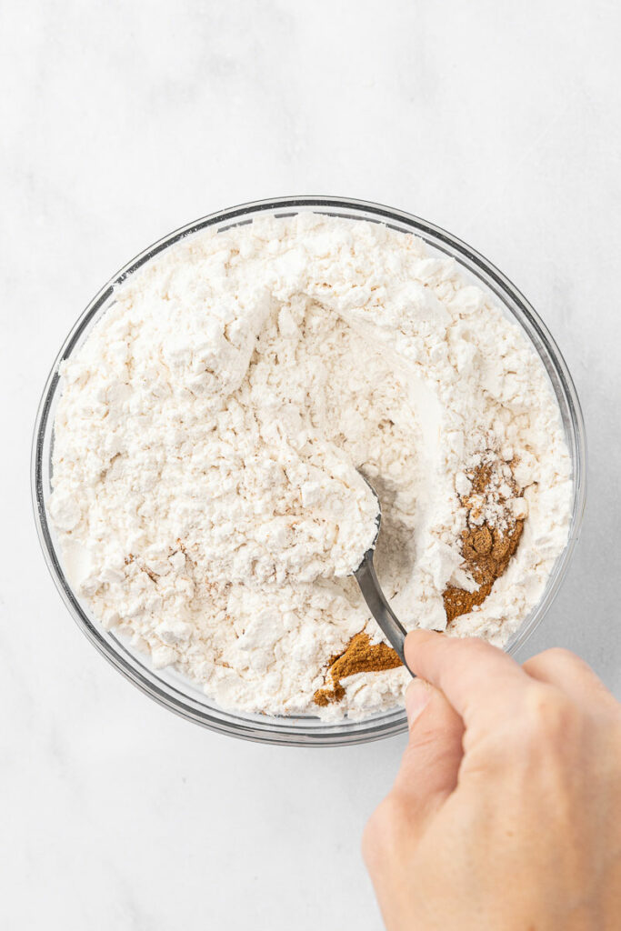 A hand holding a spoon mixing together flour, salt, baking soda, baking powder, cinnamon, and nutmeg for zucchini blueberry bread.
