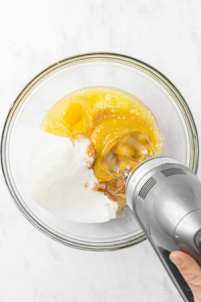 Electric mixer blending together eggs, vanilla, melted butter, and sugar in a bowl.
