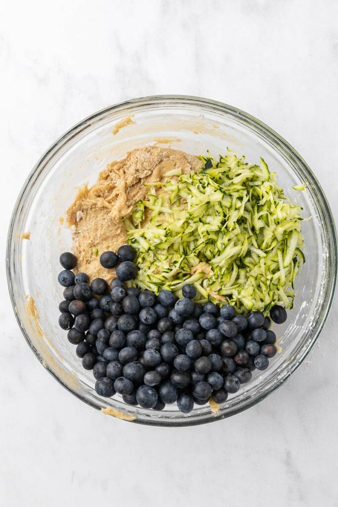Grated zucchini and whole blueberries on top of muffin batter ready to be mixed together.