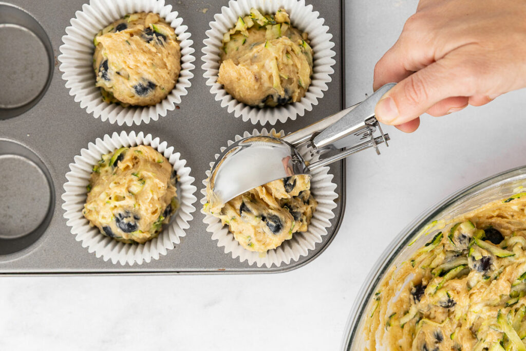 Hands using a scoop to transfer muffin batter into a muffin tin.