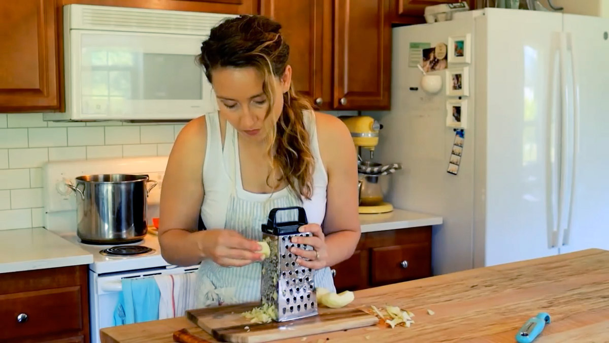 A woman using a box grater to grate a cored and peeled apple.