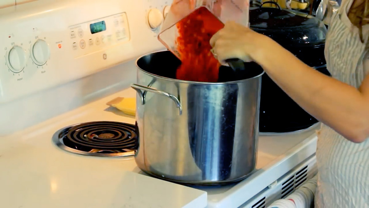 Hands pouring blended strawberries from a mixer into a large pot on a stove.