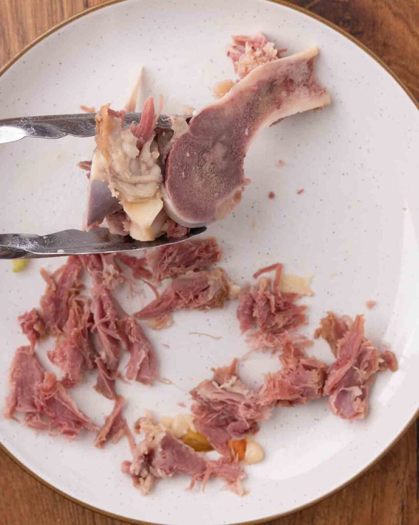 Tongs removing meat from a simmered ham bone.