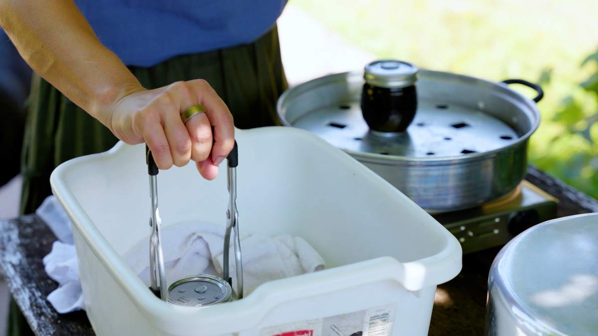 A woman moving jars of blueberry jam to a towel lined tub.