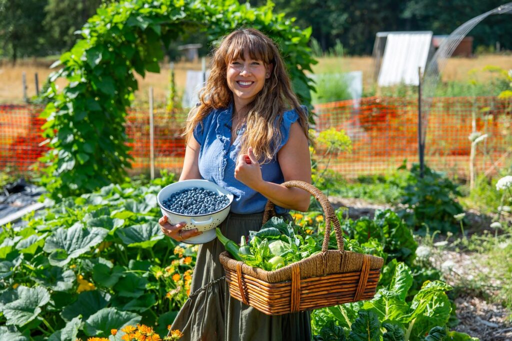A woman holding a basket of garden harvest and a large bowl of blueberries.