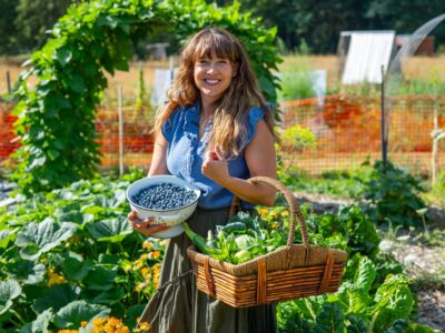 A woman holding a basket of garden harvest and a large bowl of blueberries.