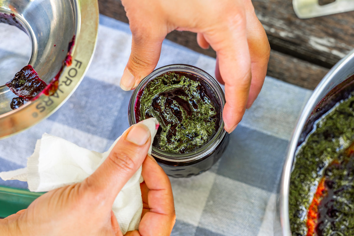 A woman's hands wiping the rim of a jar of blueberry jam.