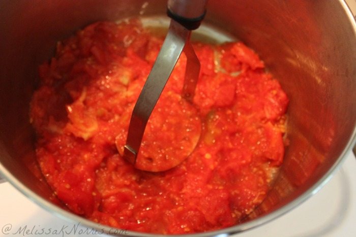 Tomatoes on the bottom of a large stockpot and a potato masher mashing the tomatoes.