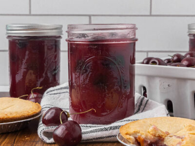 An open jar of cherry pie filling sitting amongst fresh cherries and cherry pies.