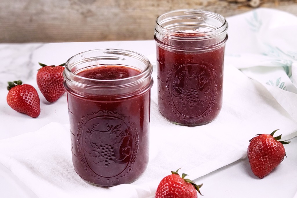 Two canning jars filled with homemade strawberry jam with strawberries on the table around it.