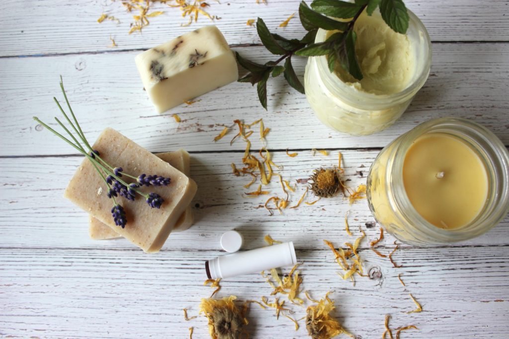 Three bars of homemade soap with dried flowers on the counter, plus a homemade candle in a mason jar, homemade body butter in a mason jar and homemade lip balm. 