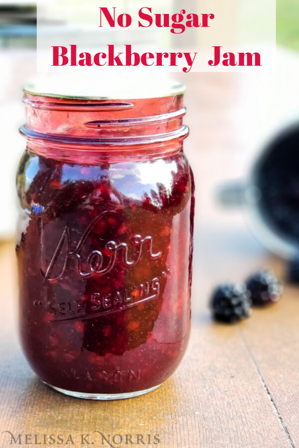 Jar of blackberry jam sitting on a table with blackberries in the background. Text overlay says, "No Sugar Blackberry Jam"