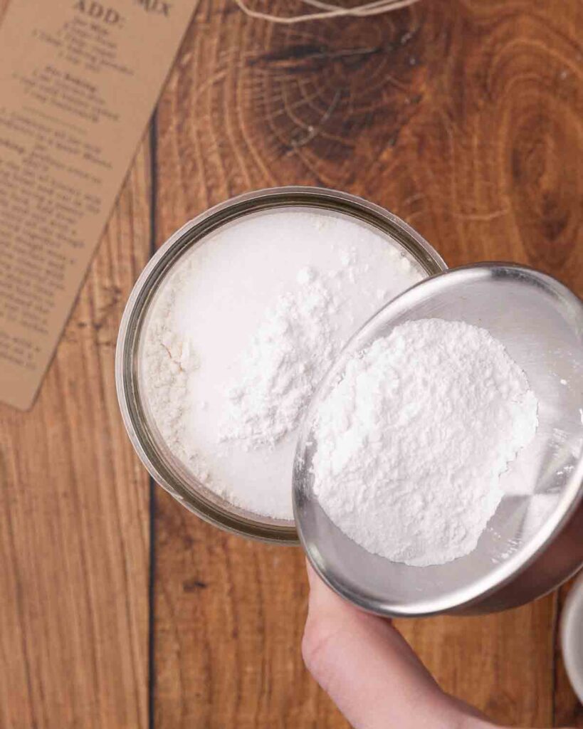 Pouring baking powder into a jar filled with biscuit mix ingredients.