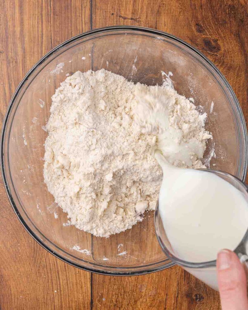 Buttermilk pouring into a glass bowl filled with dry ingredients for biscuits.