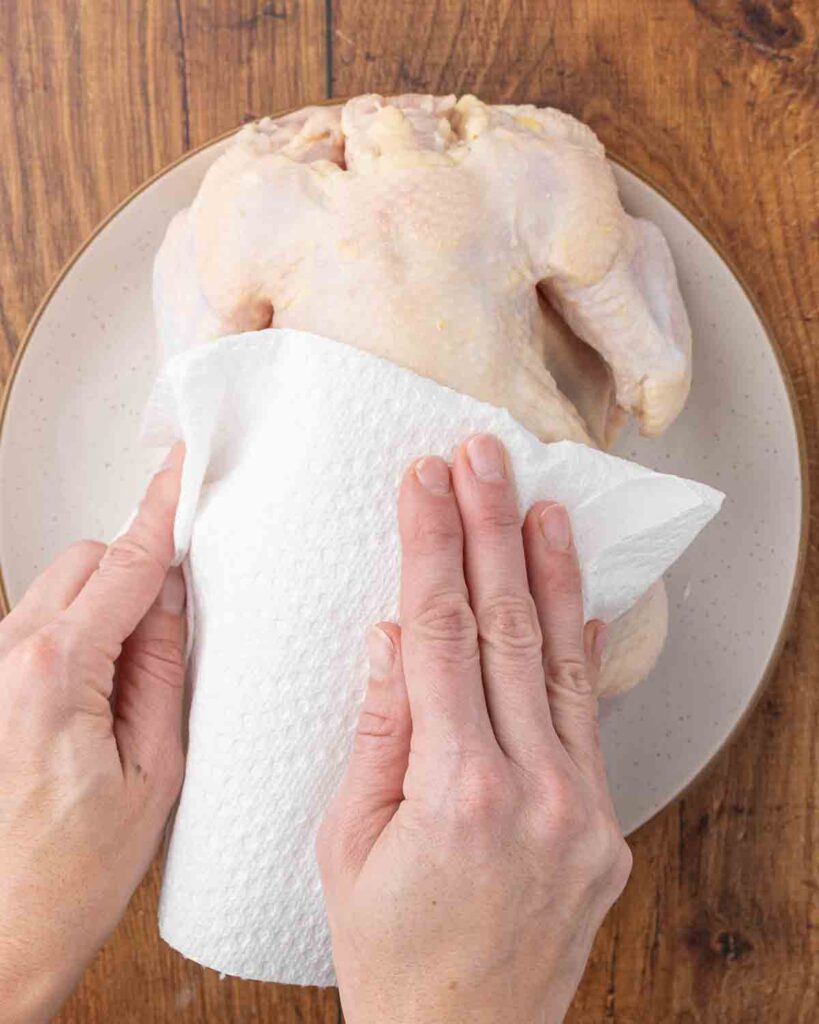Hands using paper towels to pat dry a whole raw chicken.