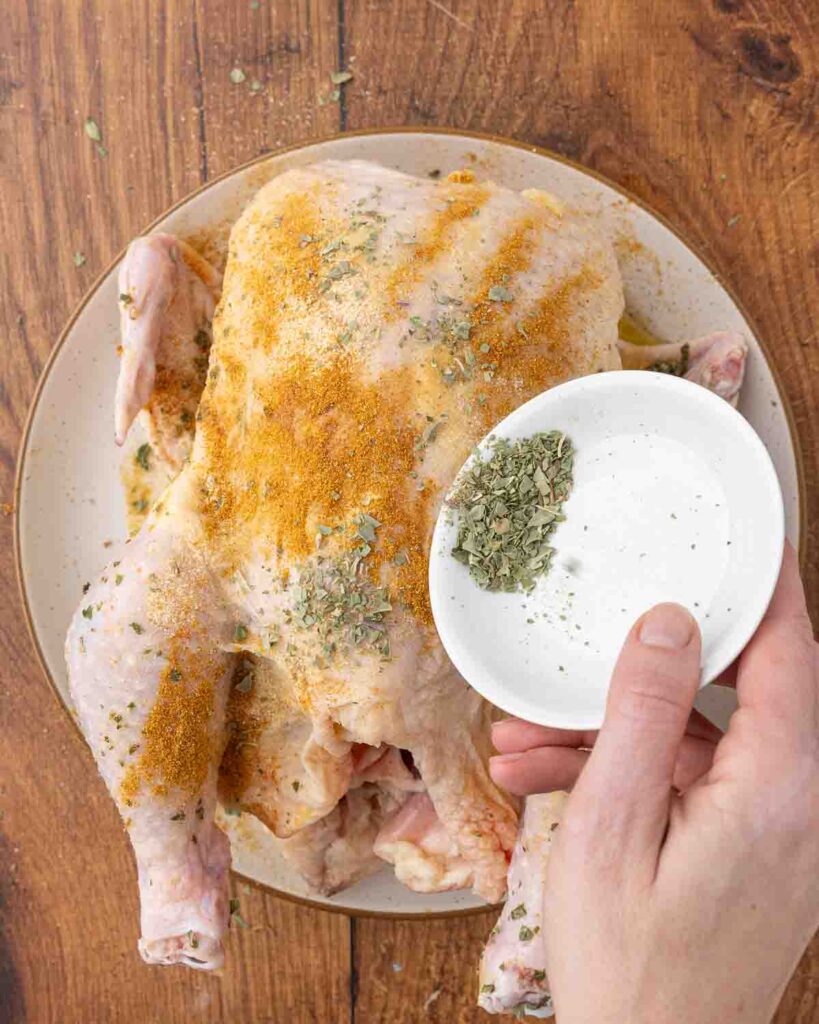 Hands sprinkling Italian seasoning, garlic powder, paprika, onion powder, and salt over a whole raw chicken.