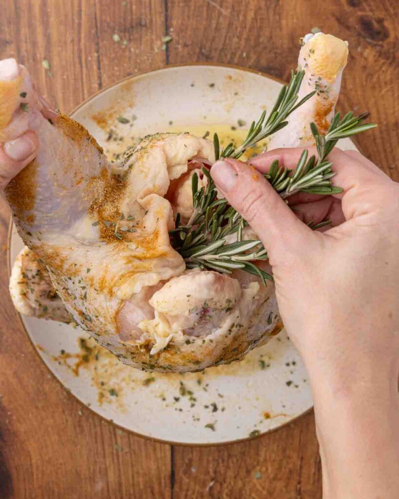 Hands placing fresh rosemary in the cavity of a whole raw chicken.