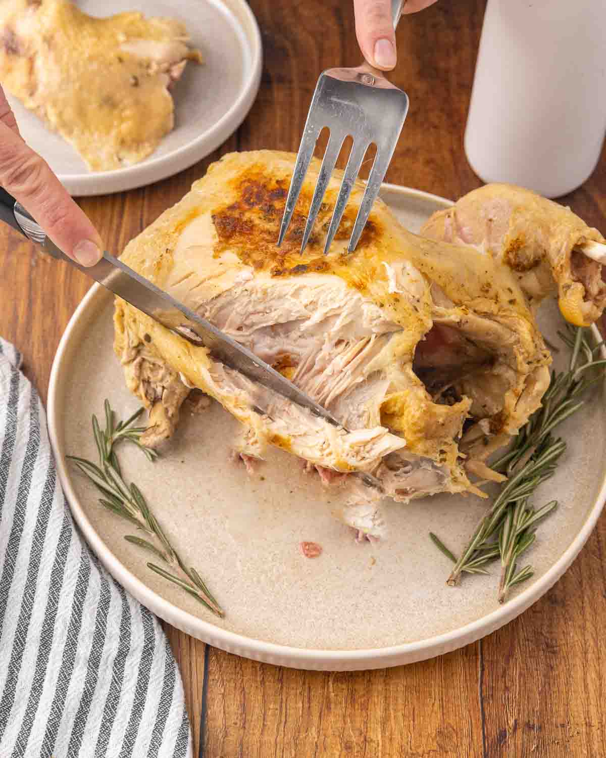 Hands holding a fork and knife carving a whole chicken on a white platter.
