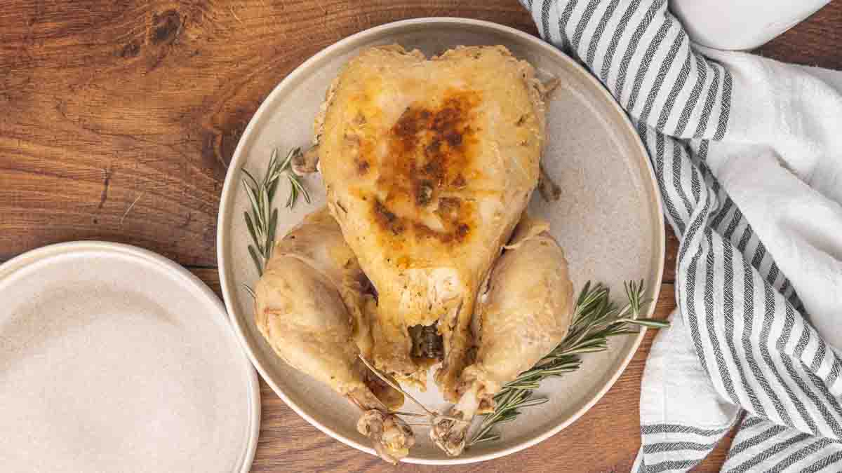 A cooked whole chicken served on a white platter and garnished with fresh rosemary.