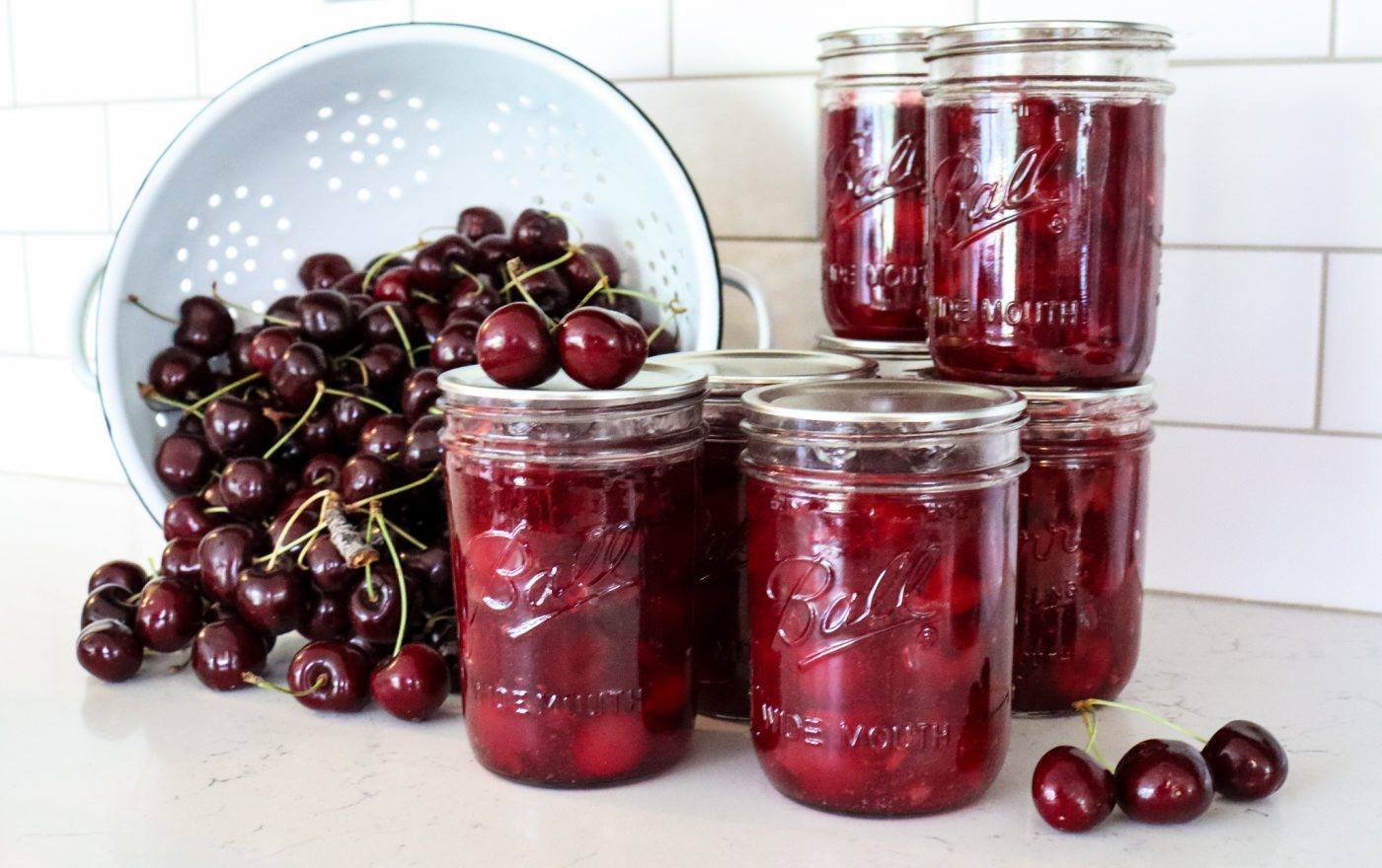 colander of cherries and jars of homemade cherry pie filling on white counter