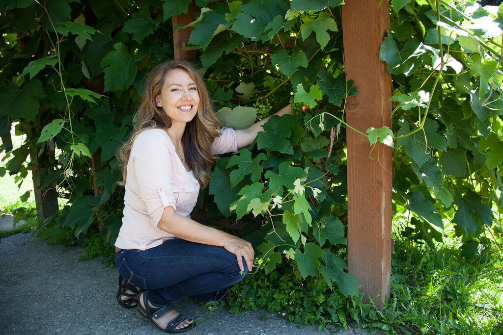 A woman kneeling beside a grape arbor full of grape vines.