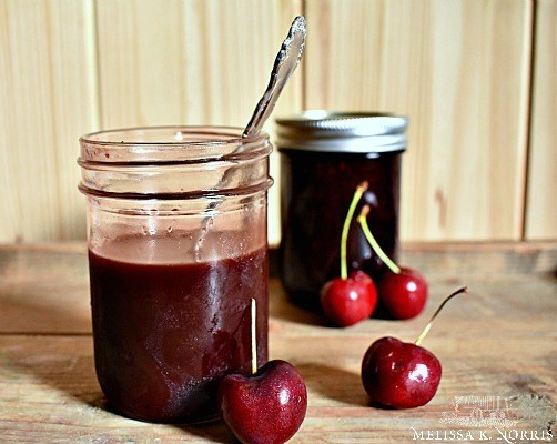 two canned jars of cherries. one opened with a spoon in it, and four cherries - all on a wooden surface