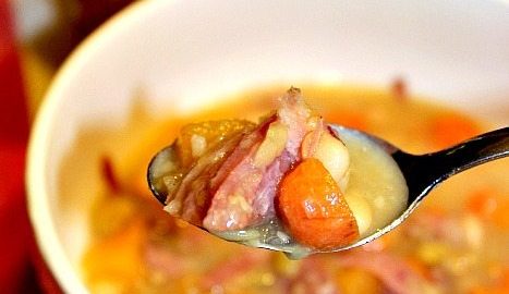 Close up image of ham and bean soup served in a white bowl with a spoon lifted to show a bite. A quart Mason Jar filled with canned bean soup sits in the background.