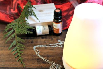 Image of an essential oil diffuser with bottles of essential oil, an essential oil diffuser necklace, a cedar bow, and red velvet draped in the background. Text overlay says, "7 Holiday Essential Oil Blends for a Natural Homestead ".