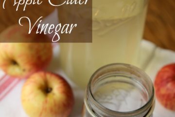 Image of a quart Mason jar filled with apple juice in the background, and a pint Mason jar filled with apple cider vinegar in forefront. Two apples are sitting to the left, and one apple is sitting to the right. Text overlay says "Easy Homemade Apple Cider Vinegar".