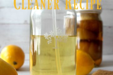 Picture of a glass spray bottle filled with vinegar cleaner in the forefront, and a Mason jar filled with lemons and vinegar in the background. Fresh lemons are scattered in between. Text overlay says "Natural Vinegar Cleaner Recipe".