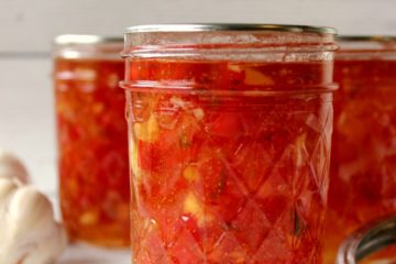Image of a jelly jar filled with red pepper jelly in the foreground. Two more jelly filled jars are in the background. A head of garlic and canning lids sit to the sides. Tex overlay says. "Red Pepper Jelly".
