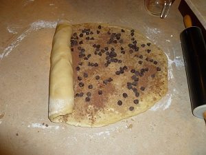 Cinnamon roll dough sprinkled with cinnamon, sugar and chocolate chips being rolled into a log.