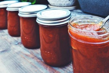 Image of three 8 ounce Mason Jars filled with spicy peach jam, stacked in a pyramid, and sitting on a whitewashed table. Placed in front of the jars is an additional opened 8 ounce Mason Jar filled with spicy peach jam with a spoon for serving. Text overlay say