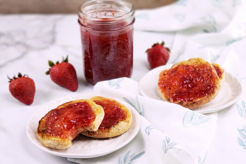 A jar of strawberry jam and jam on an english muffin.
