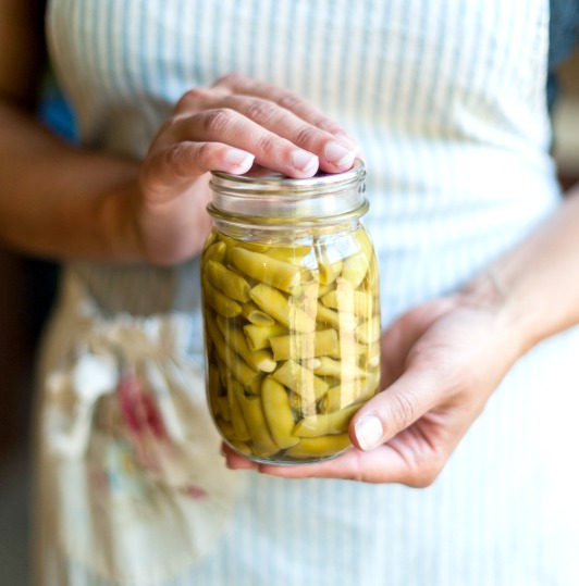 Woman in an apron holding a canned jar of green beans. Text overlay says, "How Do You Know If a Canning Recipe is Safe?"