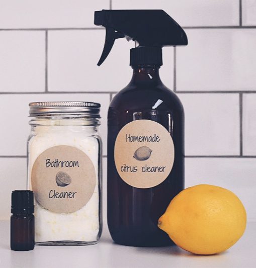 Image of an amber spray bottle and a quart Mason Jar filled with homemade cleaners against a white tile backdrop. A bottle of essential oil and a fresh lemon sit to the side. Text overlay says, "5 Homemade Natural Cleaner Recipes".