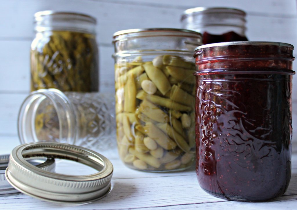 three canned jars, two of green beans and one of cherries, and an empty jar lying on its side and a canning lid on a white surface