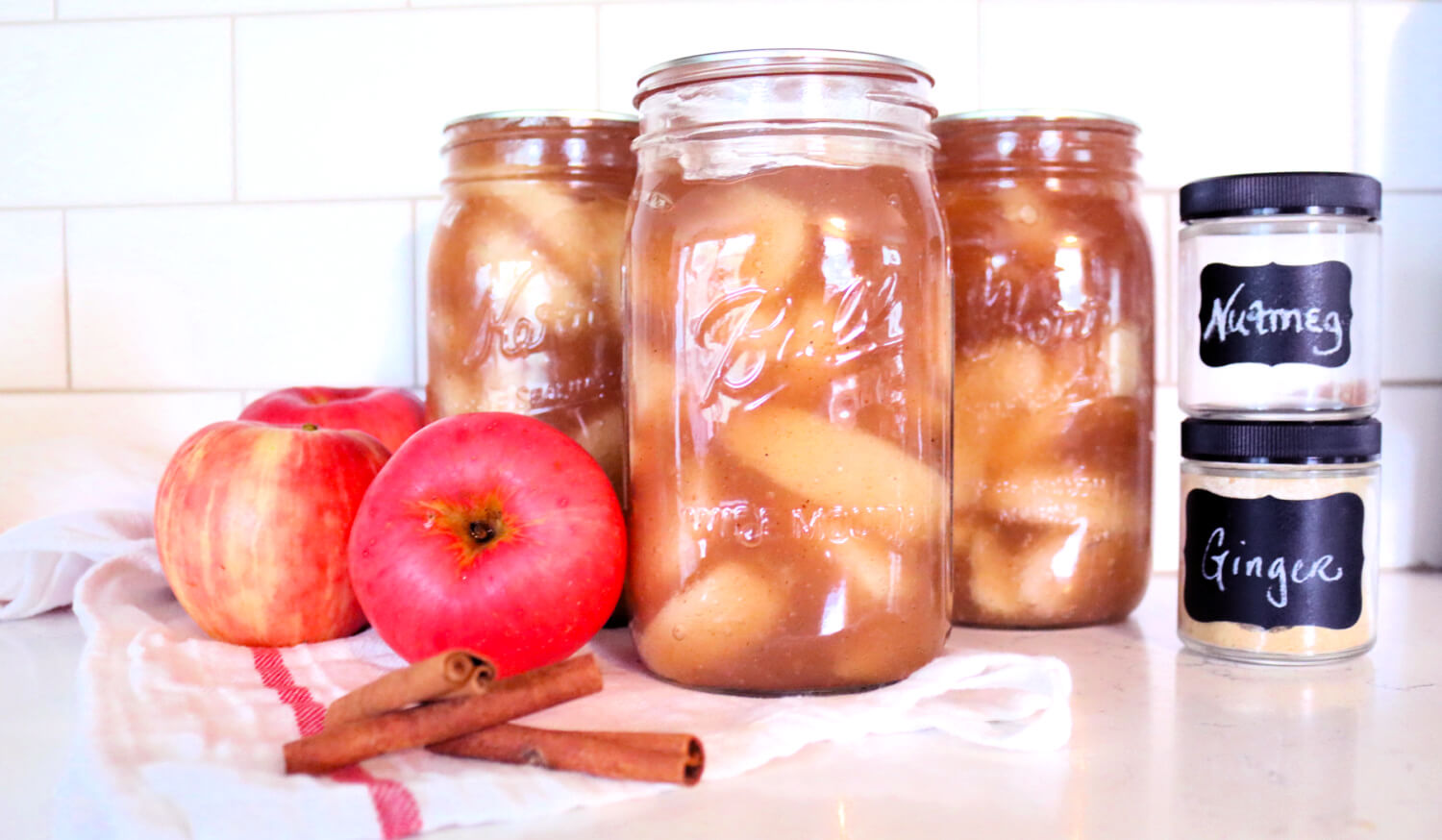 Jars of home canned apple pie filling sitting on a counter next to apples, cinnamon sticks and other spices.