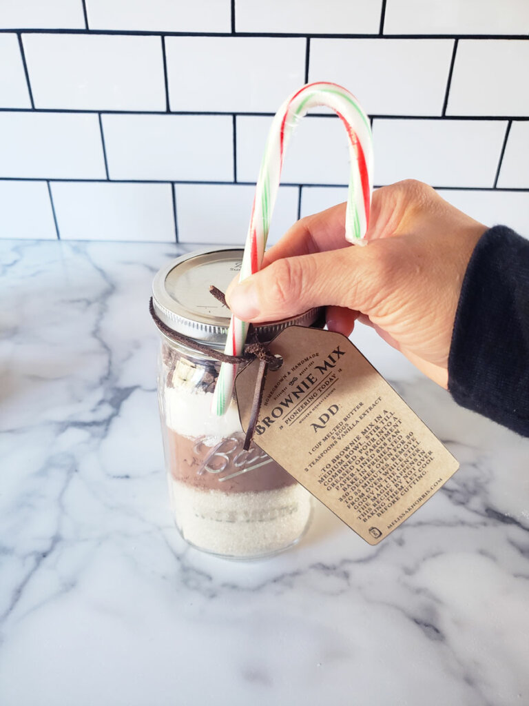 DIY Brownie mix in a mason jar with a hand attaching a candy cane to the jar.