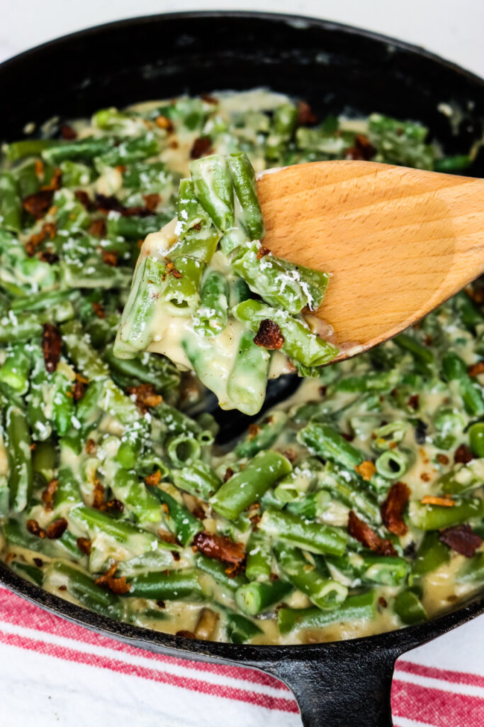 A wooden spoon taking a scoop of homemade green bean casserole from a cast iron pan.