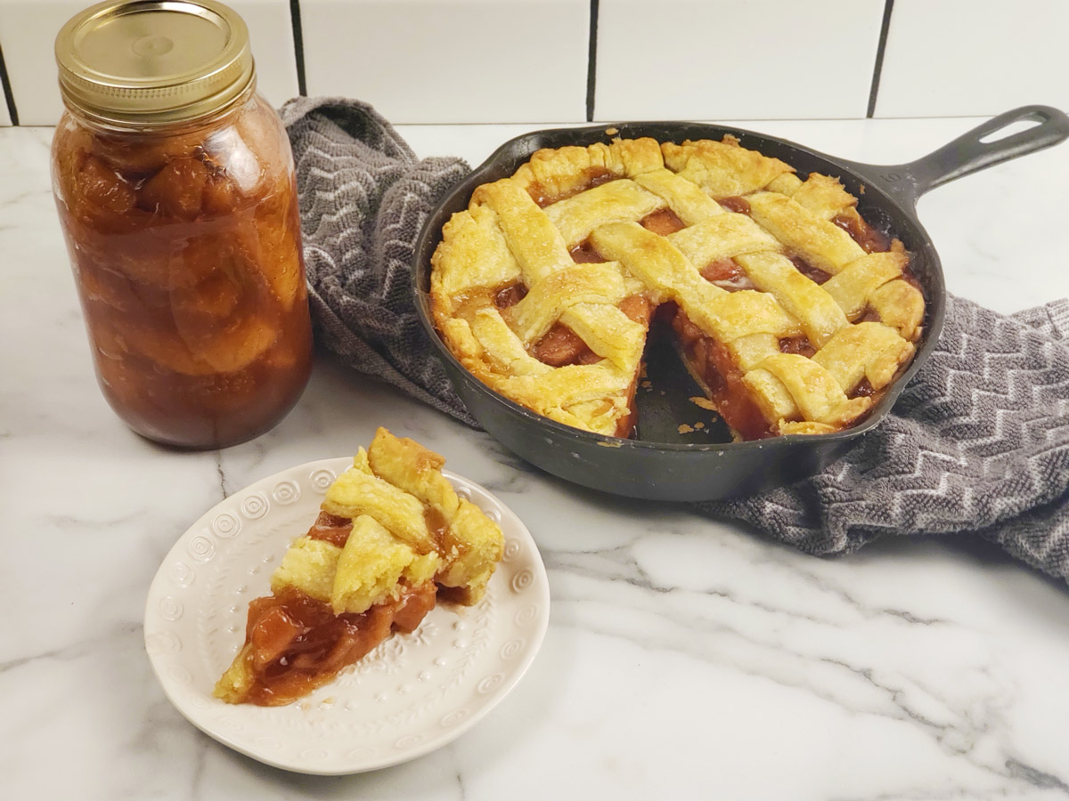 A slice of apple pie on a small white plate with a cast iron pan filled with the rest of the pie and a mason jar of apple pie filling in the background.