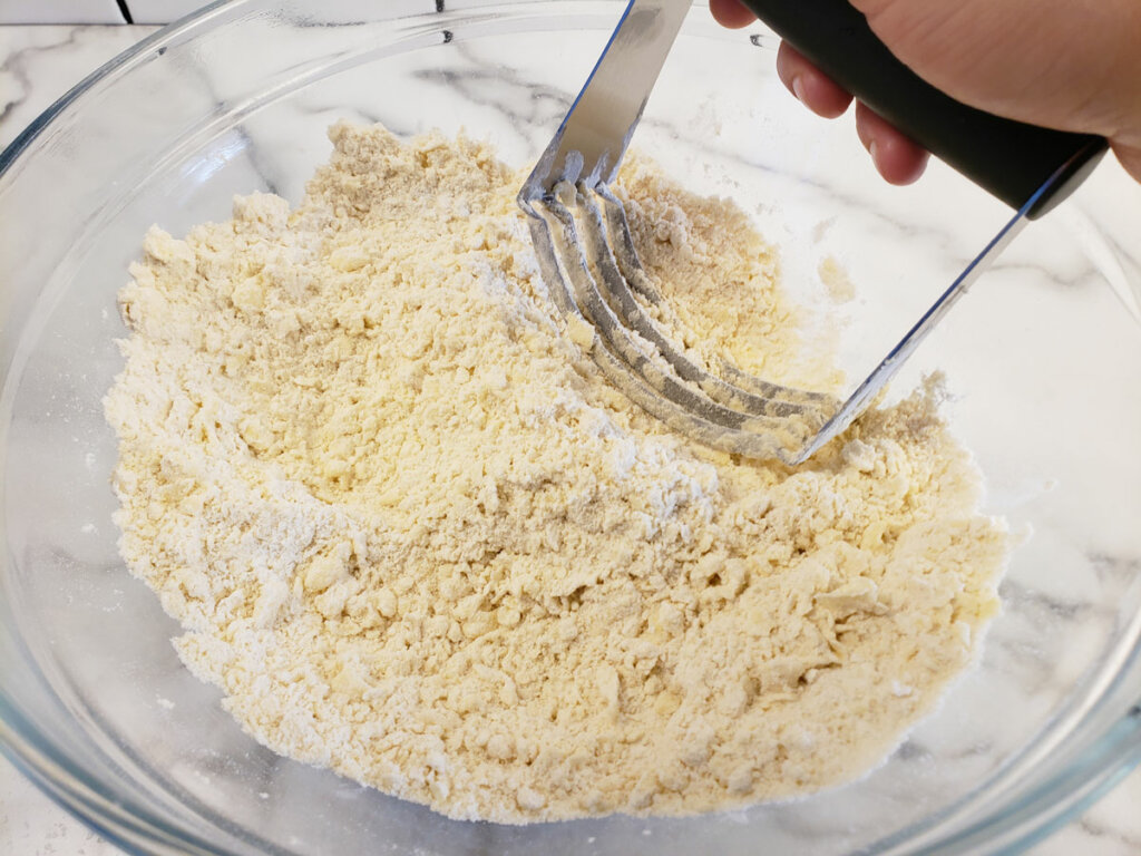A glass bowl with flour and butter and a pastry cutter cutting the butter into the flour.