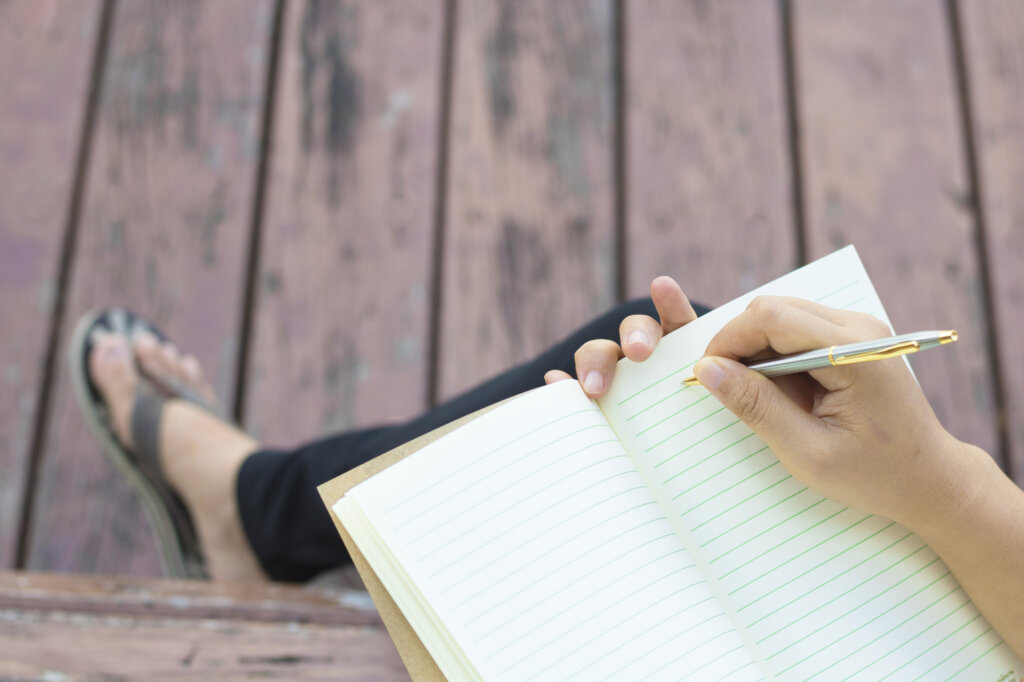 A woman writing in a notebook, sitting outside on a deck.