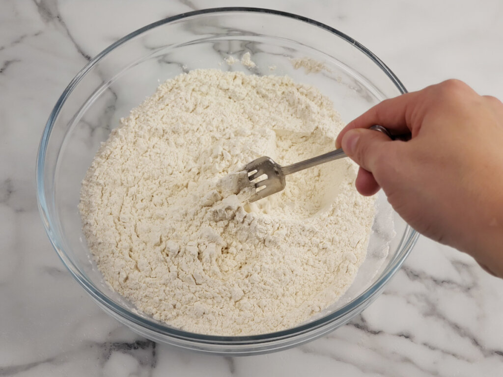 Flour and salt being stirred together in a glass bowl with a fork.