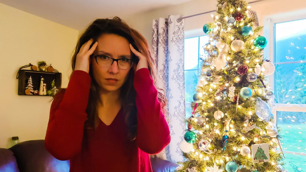 A woman holding her hands up to her head looking stressed, standing beside a Christmas tree.