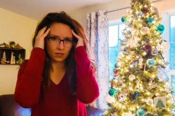 A woman holding her hands up to her head looking stressed, standing beside a Christmas tree.