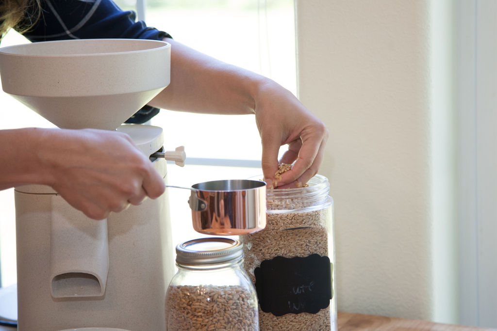 A woman's hand putting whole grain into a measuring cup with a Mockmill home grain mill in the background.