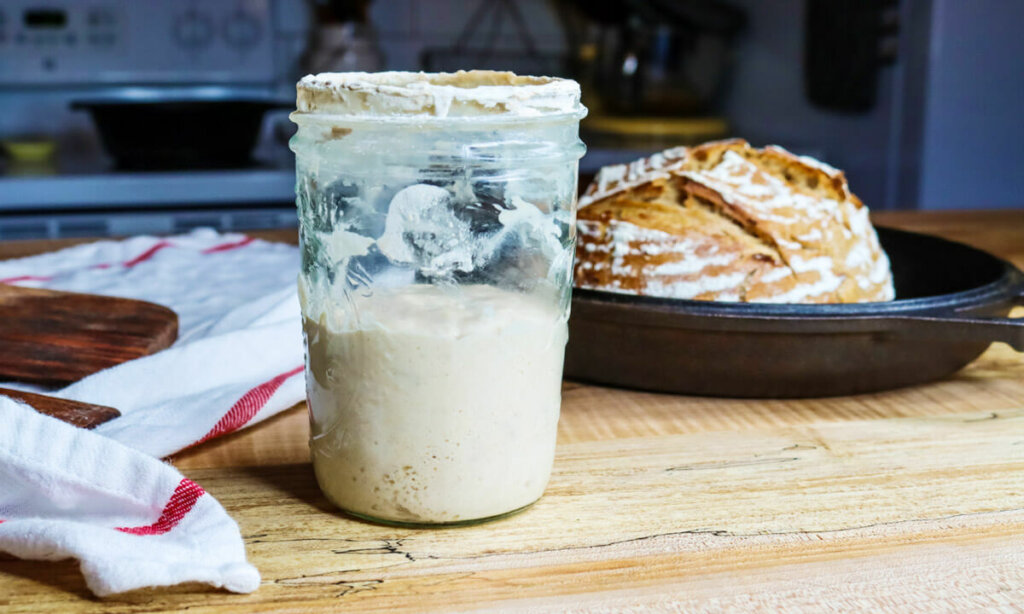 Sourdough starter in a jar with a loaf of bread in the background.