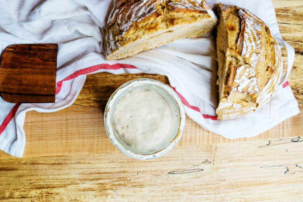 Vertical shot of a loaf of bread with a slice cut out and a jar of sourdough starter.