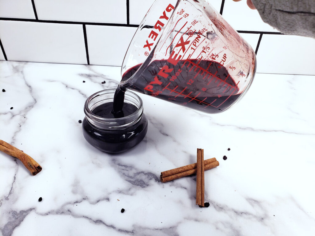 Elderberry syrup being poured from a measuring cup into a small glass jar.